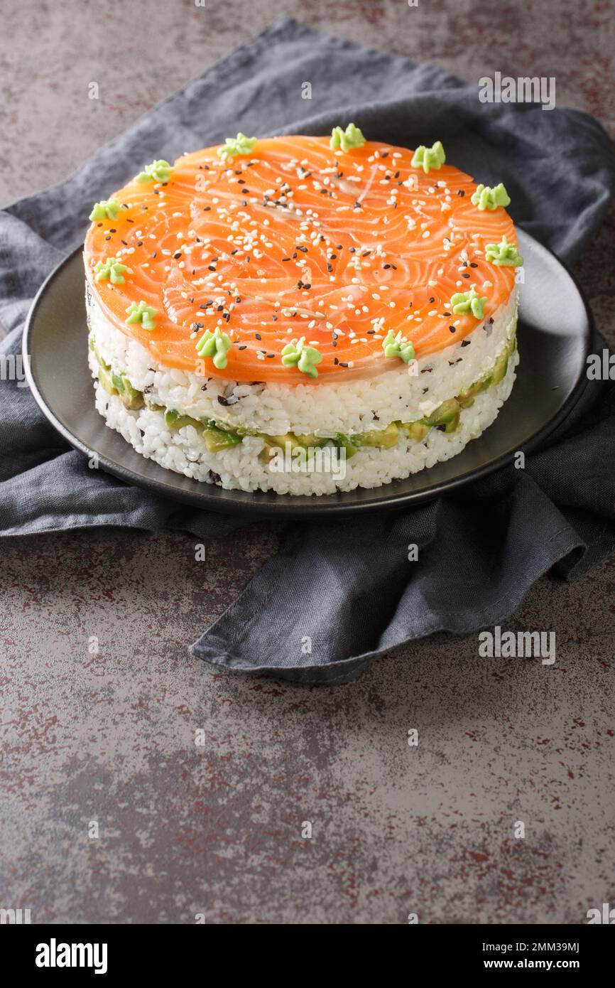 Lachs-Sushi-Kuchen mit Reis, Avocado, Frischkäse, Seetang, Sesamsamen, dekoriert mit Wasabi Nahaufnahme auf einem Teller auf dem Tisch. Vertikal Stockfoto