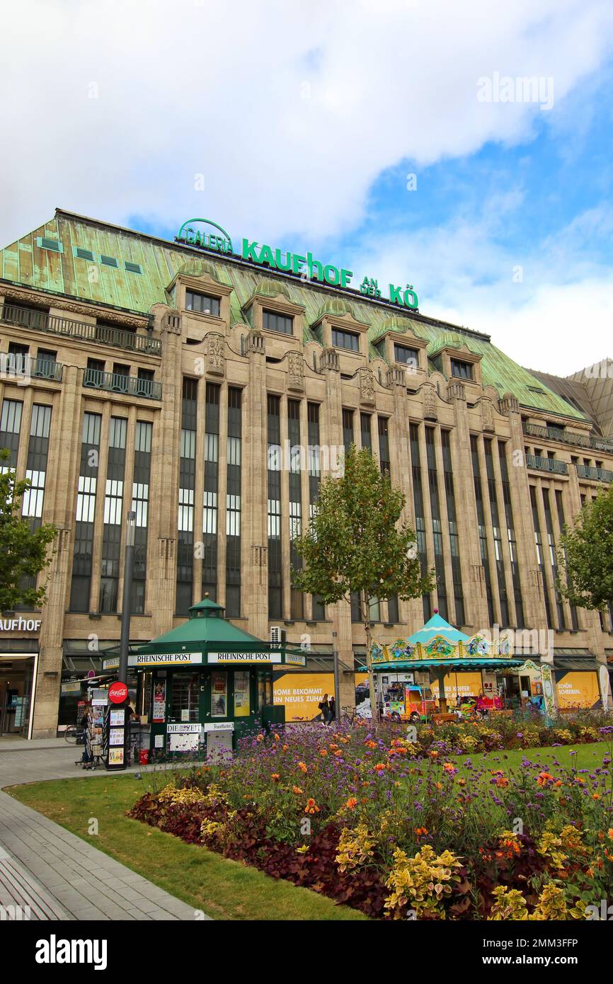 Weitwinkelblick auf das historische Gebäude „Kaufhof an der Kö“ mit Zeitungskiosk und Karussell vor dem Hotel. Galeria Kaufhof ist eine deutsche Kaufhauskette. Stockfoto