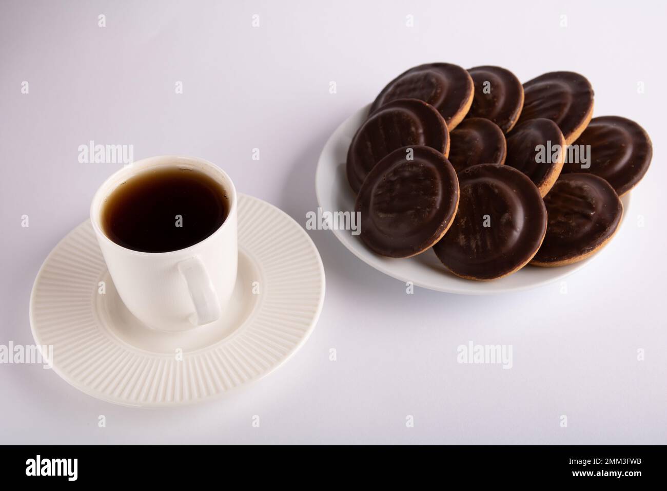 Fotografieren Sie eine Kaffeetasse mit Kaffeeständen neben einem vollen Teller mit Schokoladen überzogenen Keksen auf weißem Hintergrund von oben Stockfoto