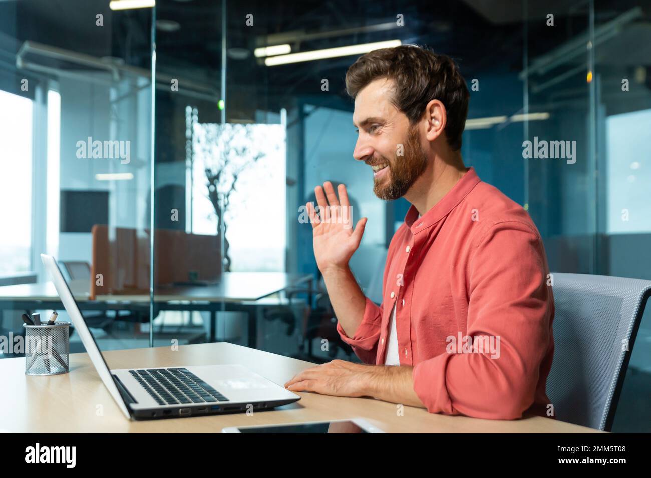 Porträt eines jungen Mannes in einem roten Hemd. Im Büro am Tisch sitzen, an einem Laptop arbeiten, per Videoanruf kommunizieren, winken, lächeln. Stockfoto