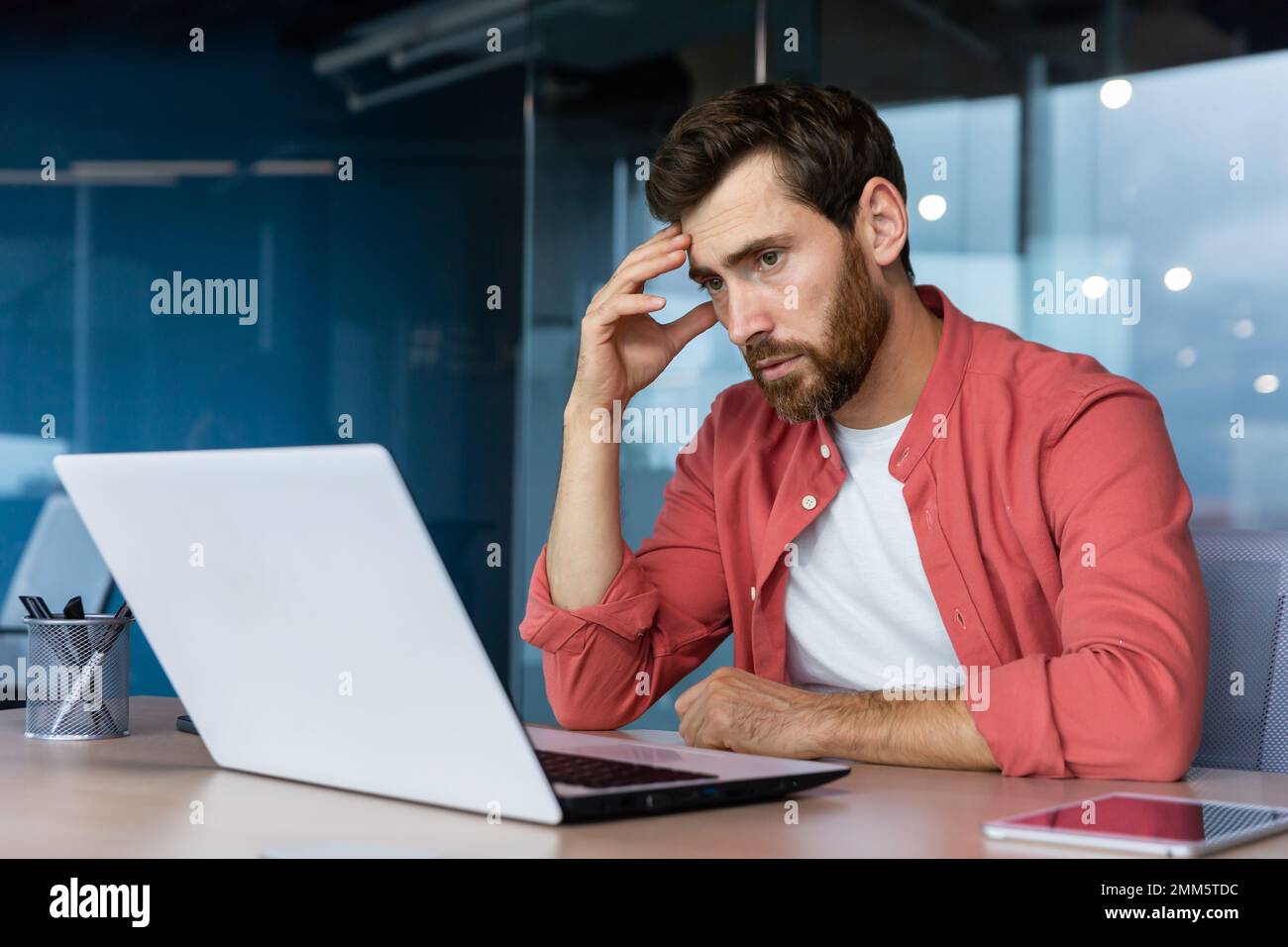 Ein gestresster und müder junger Geschäftsmann, Student, Freiberufler in einem roten Hemd sitzt im Büro an einem Laptop. Er hält seinen Kopf. Überstunden, Deadline, Probleme. Stockfoto