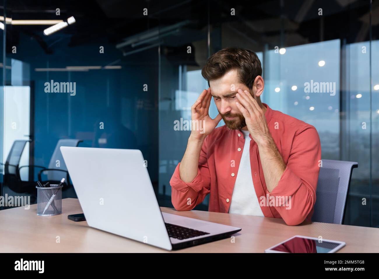 Stress und Kopfschmerzen am Arbeitsplatz. Ein junger Geschäftsmann in einem roten Hemd sitzt am Schreibtisch im Büro, hält seinen Kopf und schließt die Augen. Er fühlt sich schlecht, macht sich Sorgen um Probleme, ein Projekt, Überstunden, Stockfoto