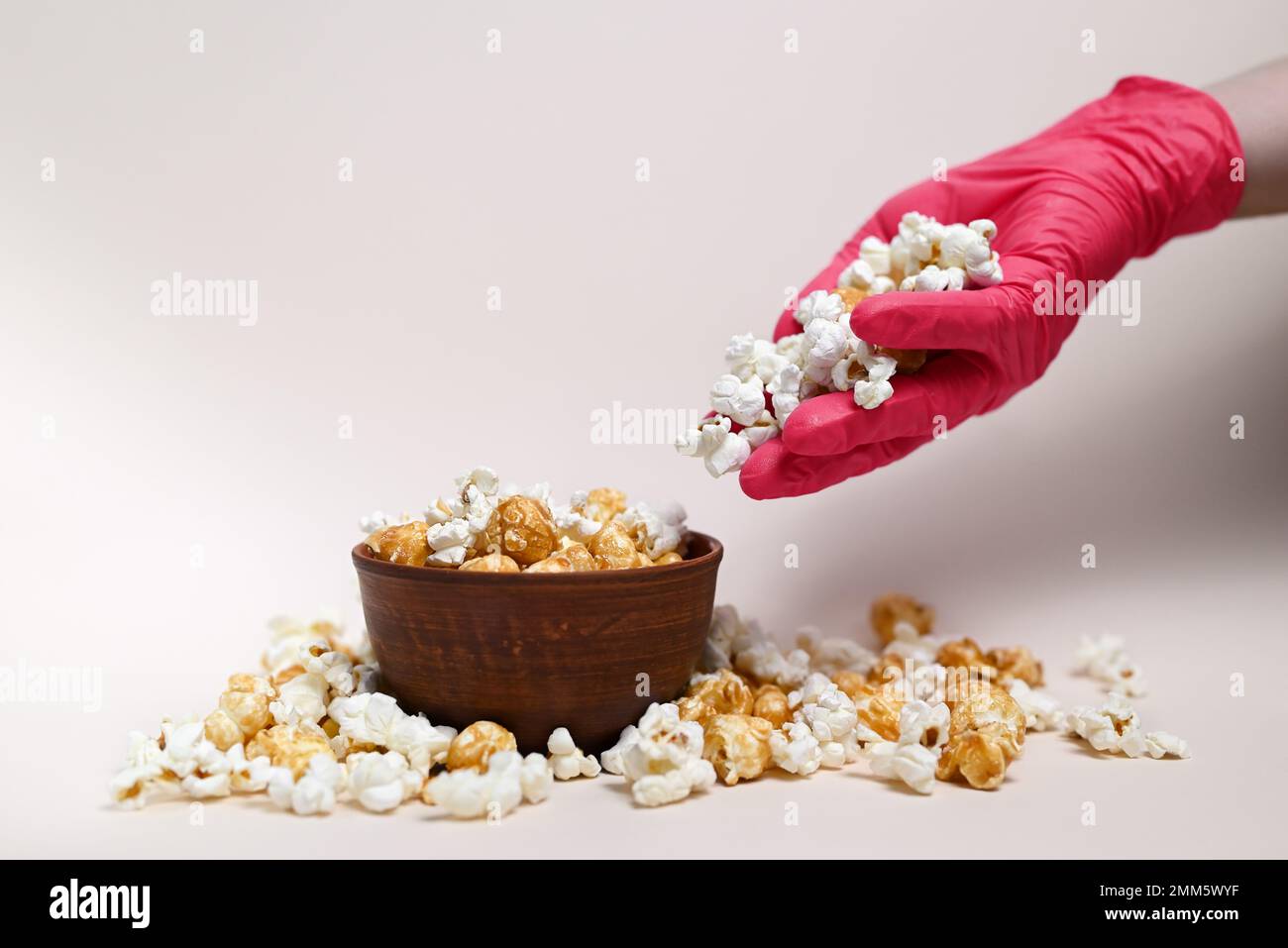Abstrakte Zusammensetzung. Popcorn-Mix in einer Keramikschüssel. Popcorn läuft von der Hand in die Schüssel. Auf der Hand ist eine medizinische Handschuhfarbe Viva Magenta. Stockfoto