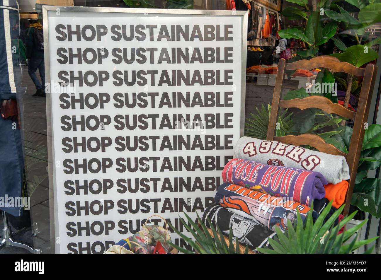 Ein Schaufenster mit einem Schild mit der Aufschrift „Shop Sustainable“ und mehreren gestapelten Sweatshirts auf einem Stuhl – umweltfreundliche und sozial verantwortliche Mode. Stockfoto