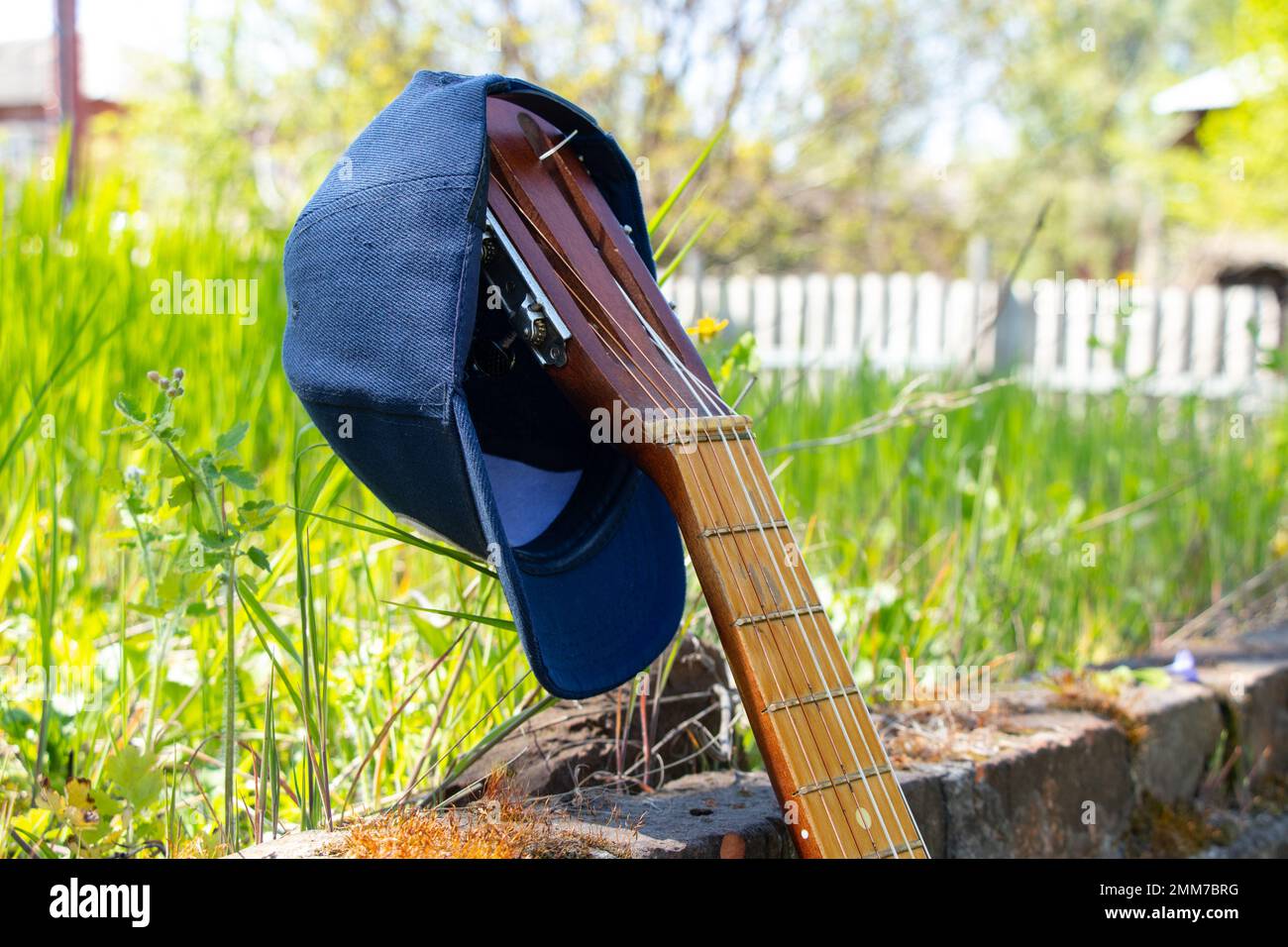 Baseballmütze, die an einer alten Gitarre hängt, steht auf einem Hintergrund aus Gras Stockfoto