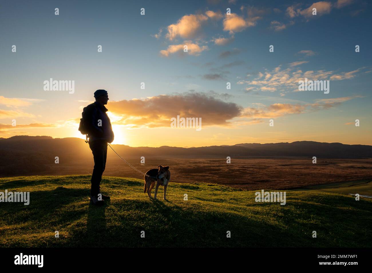 Person mit einem Hund auf dem Gipfel der Dunadd Hill Fort bei Sonnenuntergang, in der Nähe von Lochgilphead, Argyll, Schottland, Großbritannien Stockfoto