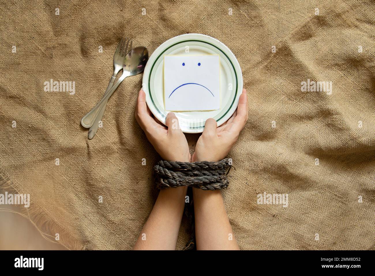 Ein Teller mit Papier, auf dem ein trauriges Smiley gezeichnet wird und die Hände der Frauen mit einem Seil gefesselt sind, Hunger und nichts zu essen, Sklaverei Stockfoto