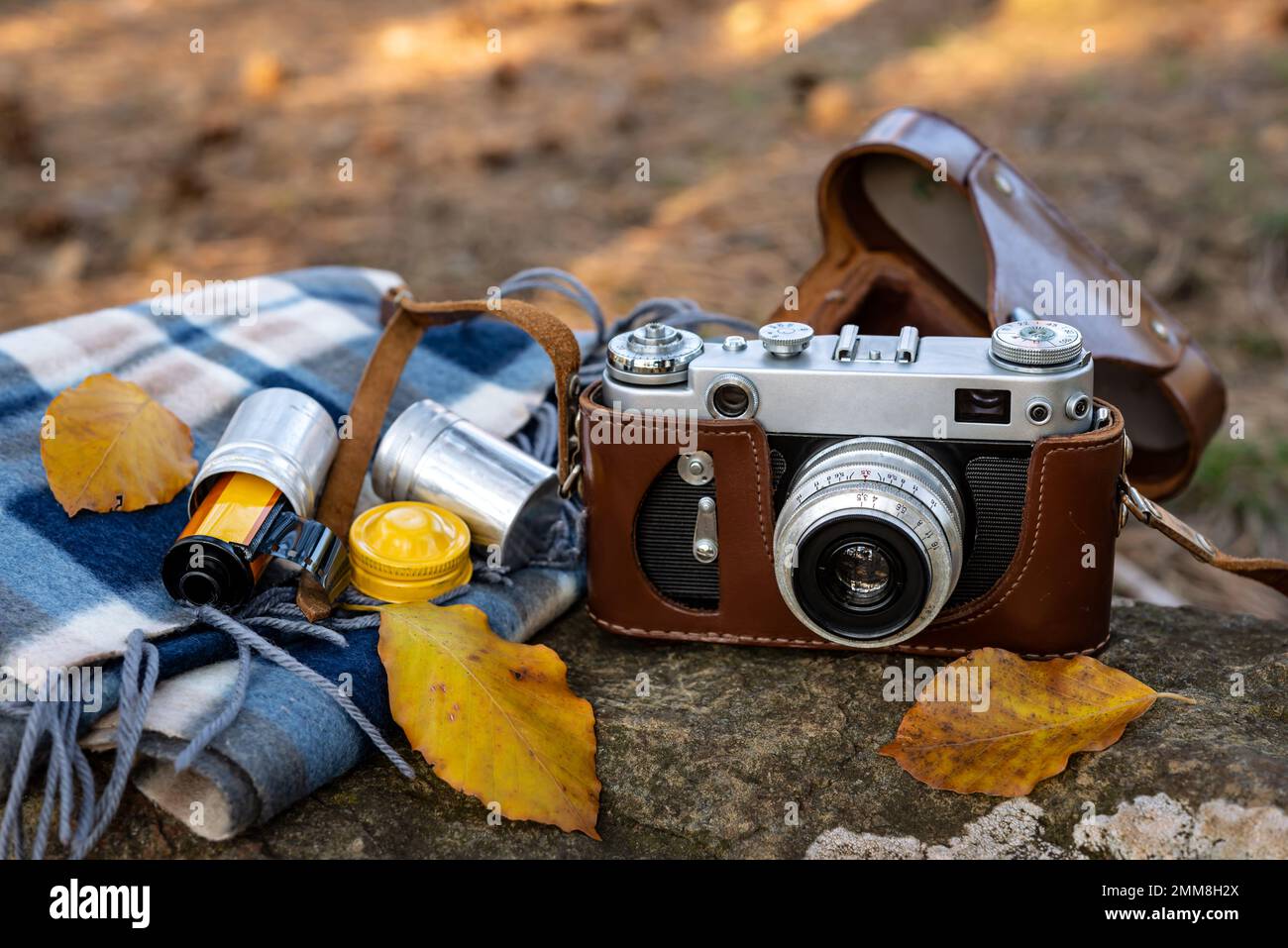 Oldtimer-Fotokamera in brauner Ledertasche und Fotofilm in einer Kartusche mit Schal und Herbstblättern auf einem Stein im Freien. Selektiver Fokus. Stockfoto