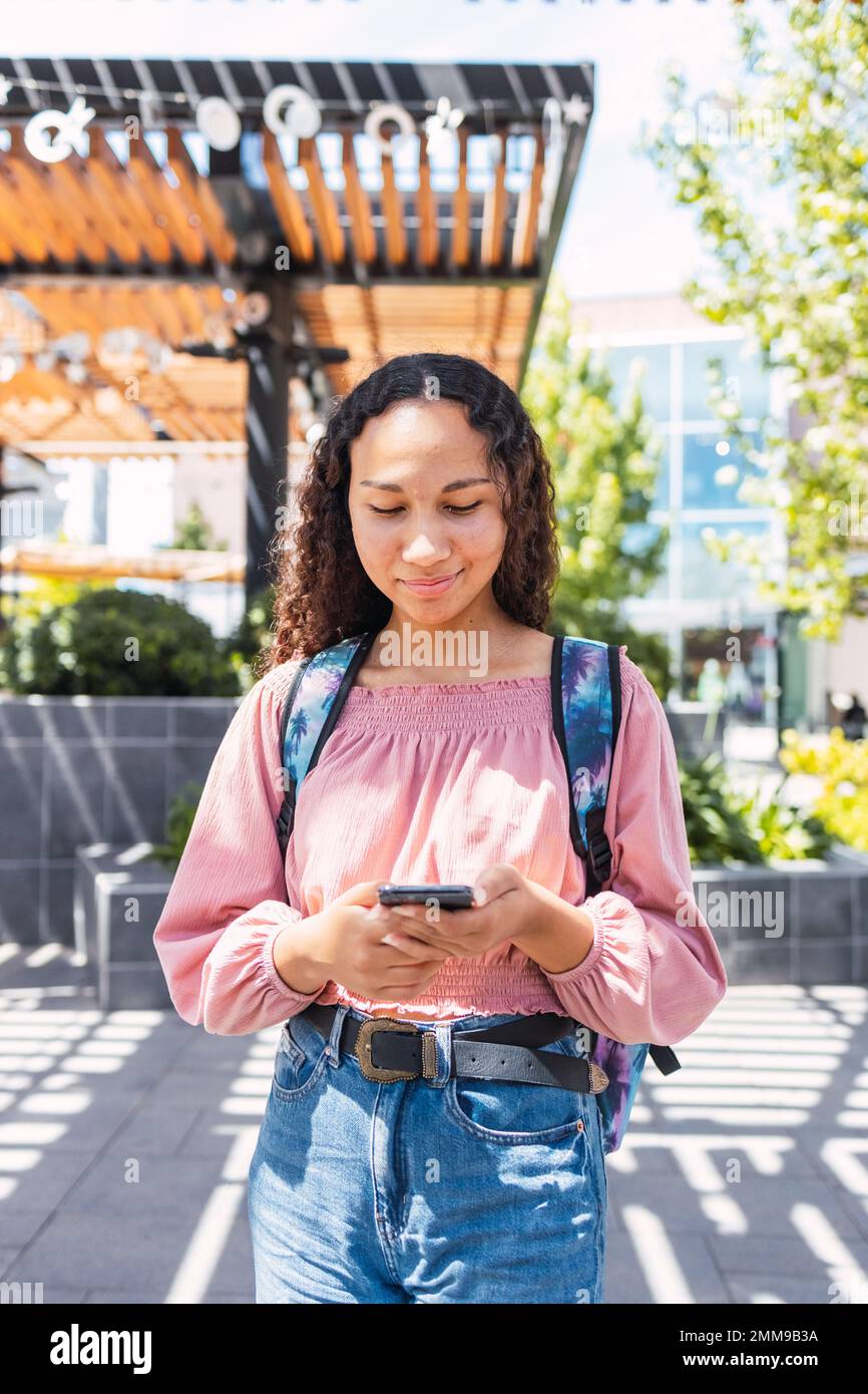 Lateinische Studentin, die lächelt und ihr Smartphone außerhalb des Einkaufszentrums in ihrer Freizeit benutzt Stockfoto