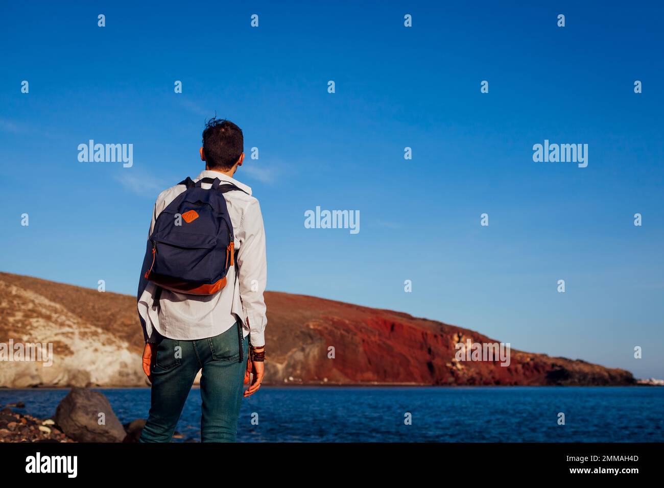 Reisender mit Rucksack, der die rote Strandlandschaft auf Santorini Island, Griechenland, mit Hügeln und Meer bewundert. Allein unterwegs. Sommerferien Stockfoto Reisender mit Rucksack, der die rote Strandlandschaft auf Santorini Island, Griechenland, mit Hügeln und Meer bewundert. Allein unterwegs. Sommerferien Stockfoto