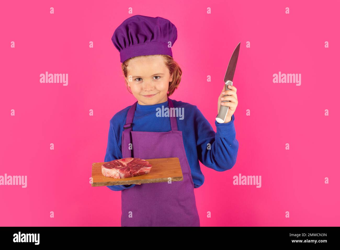 Kinderkoch hält Schneidebrett mit Fleischsteak und Messer. Teenager Junge mit Schürze und Chefhut bereitet eine gesunde Mahlzeit zu. Stockfoto