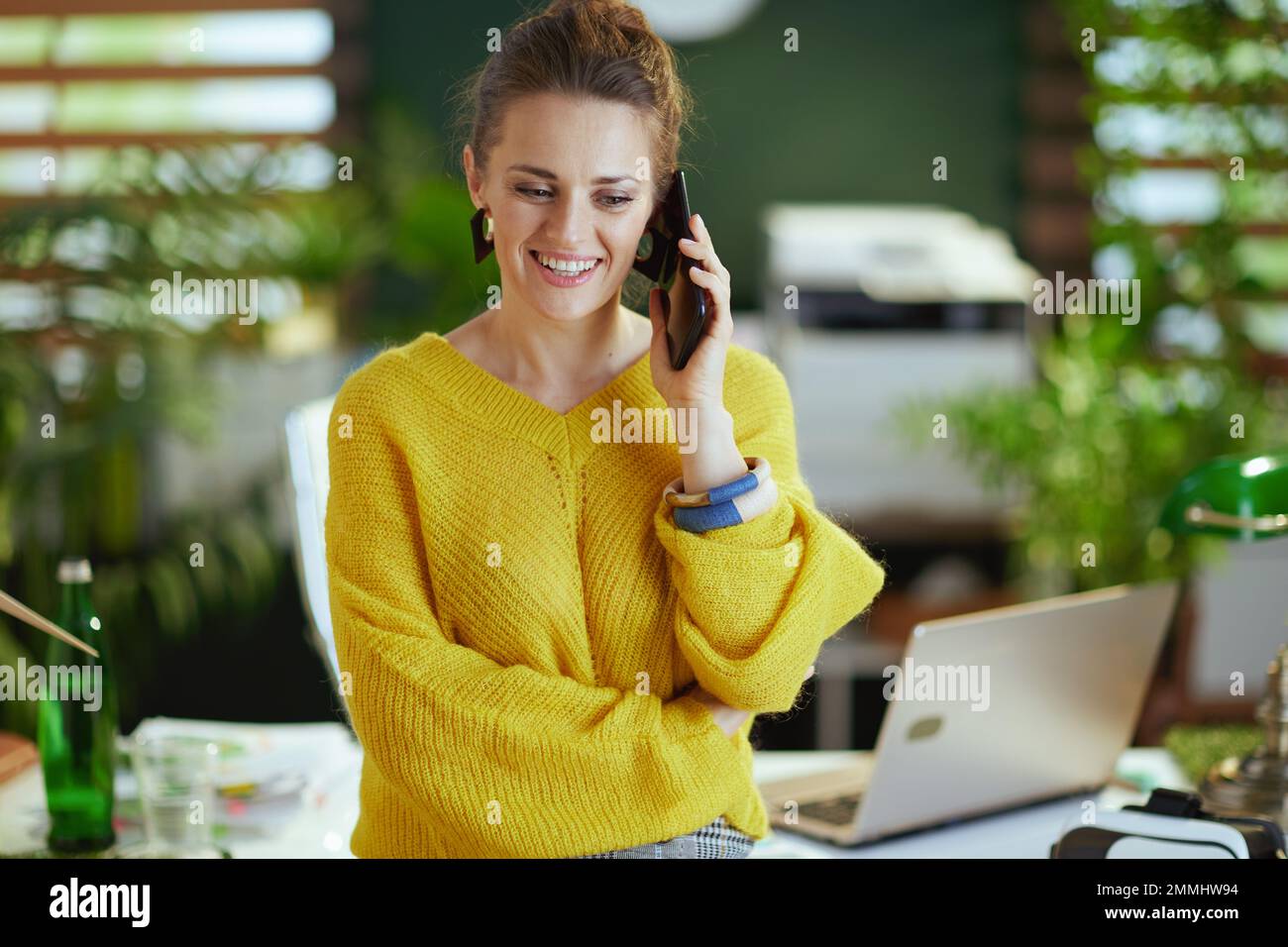 Lächelnde Eigentümerin eines kleinen Unternehmens mittleren Alters in einem gelben Pullover im modernen grünen Büro, die auf einem Smartphone spricht. Stockfoto