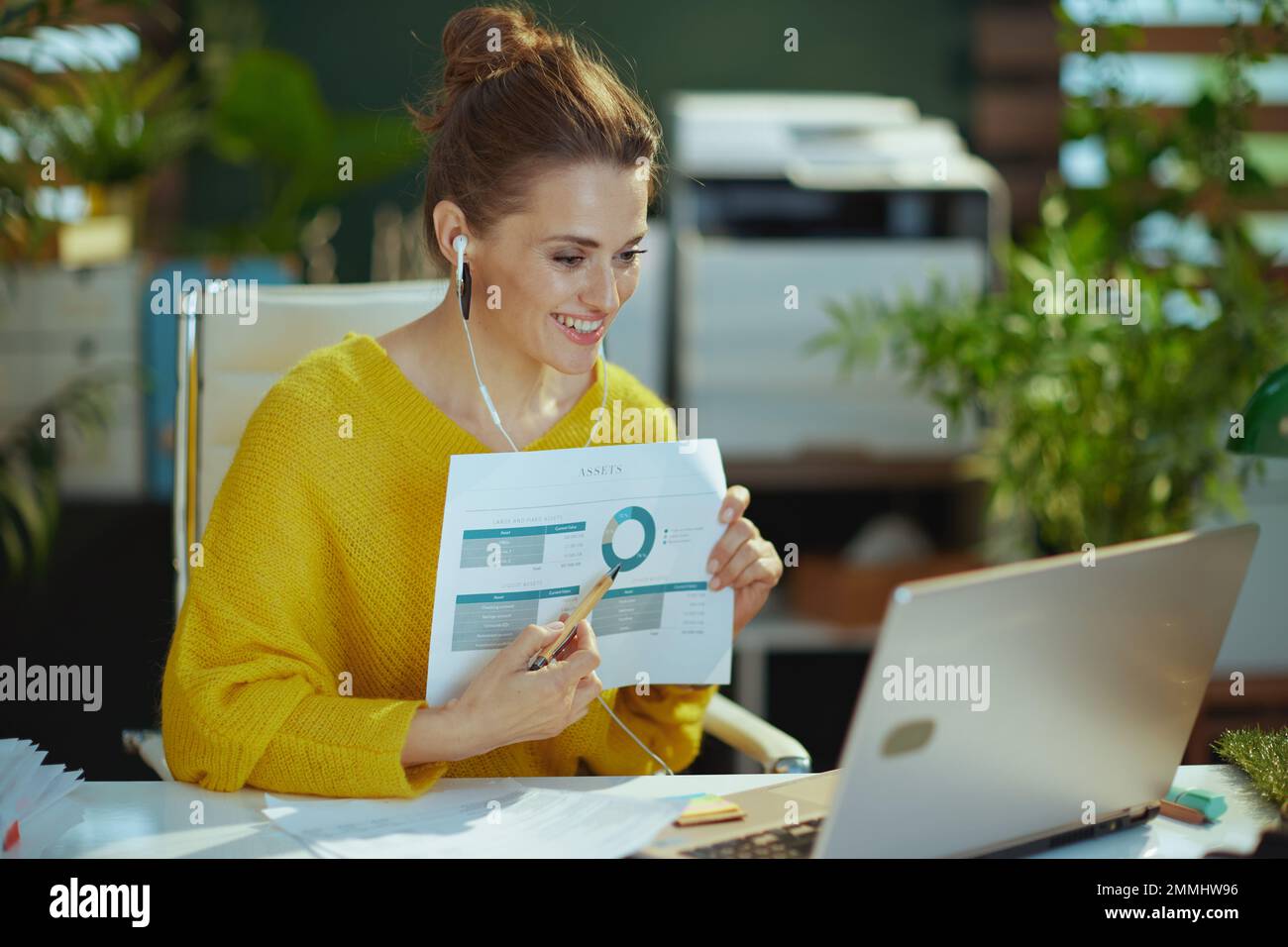 Lächelnde, trendige Eigentümerin eines kleinen Unternehmens in gelbem Pullover mit Laptop, die Videokonferenz veranstaltet und im modernen grünen Büro Karten zeigt. Stockfoto