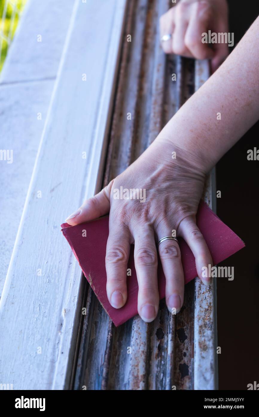 Die Hände einer Frau mit Schleifpapier, die vor dem Malen einen Fensterrahmen schleift. Das Konzept der ermächtigten Frau. Vertikale Ausrichtung. Stockfoto
