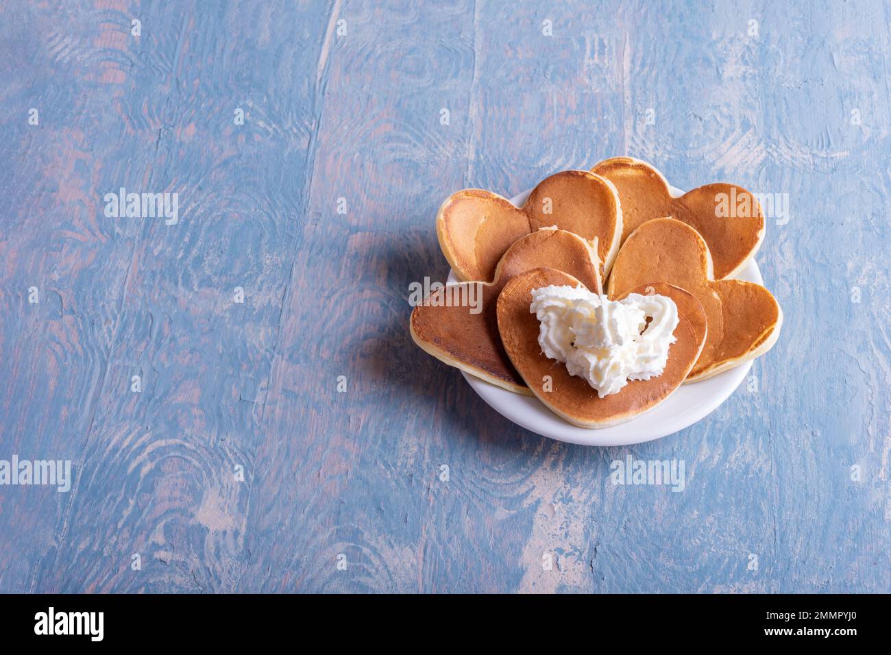 Hausgemachter herzförmiger Pfannkuchen mit weißer Creme auf einem weißen Teller auf einem blauen Holztisch, Kopierbereich, Draufsicht. Originelle, kreative Valentinstagsferien Stockfoto