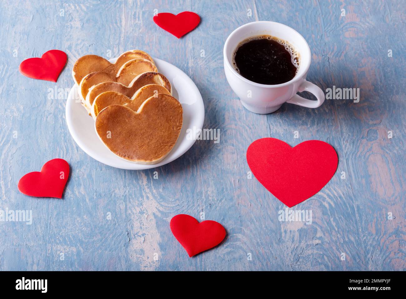 Hausgemachte Pfannkuchen in Form eines Herzens auf einem weißen Teller, eine Tasse mit Kaffee oder Kakao auf einem blauen, stilvollen Holzhintergrund mit Herzen. Frühstück für Stockfoto