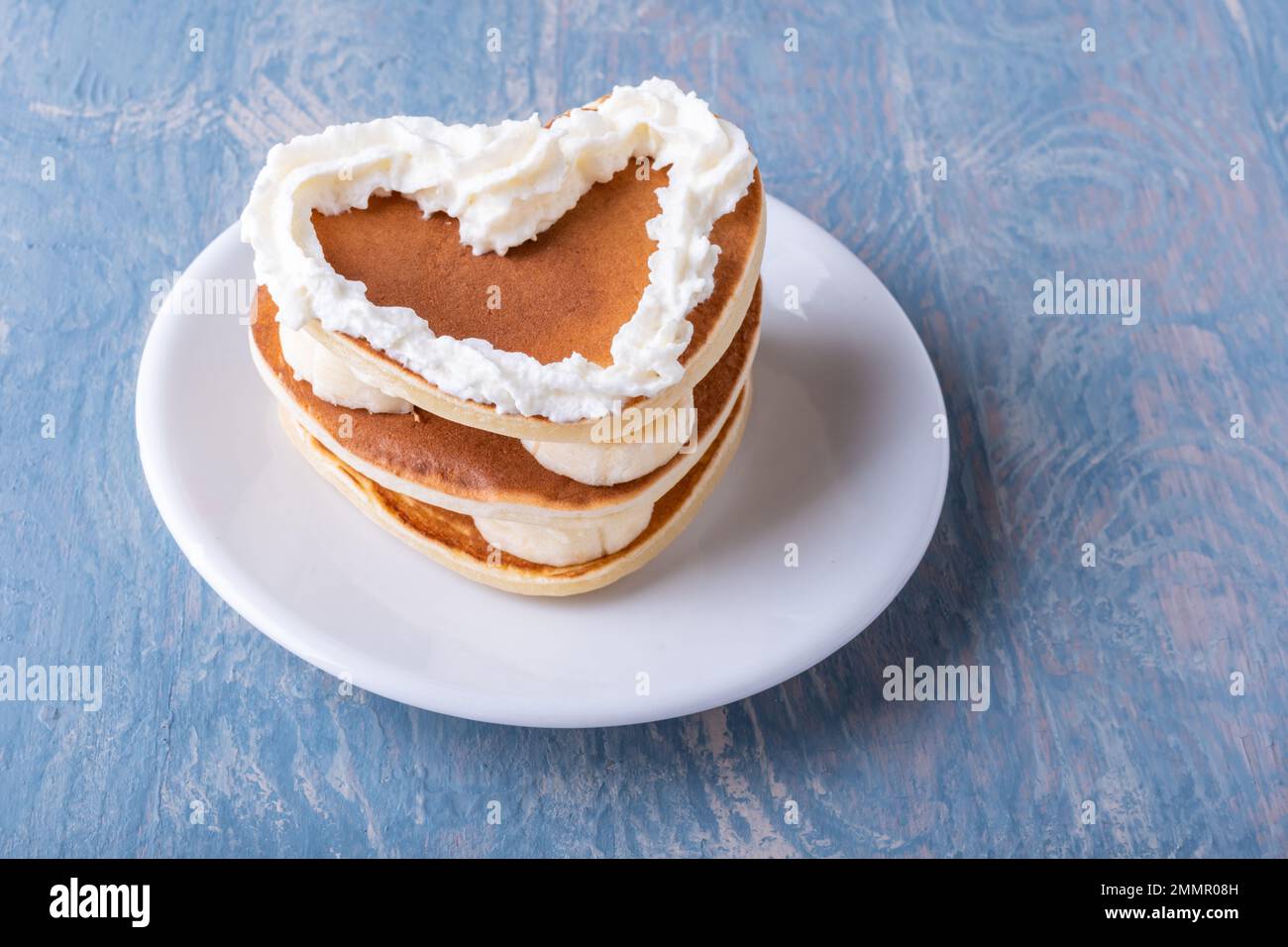 Kreatives Frühstück am Valentinstag. Hausgemachter herzförmiger Pfannkuchen mit Banane, dekoriert mit weißer Creme auf einem weißen Teller auf einem blauen Holztisch, Kopie Stockfoto