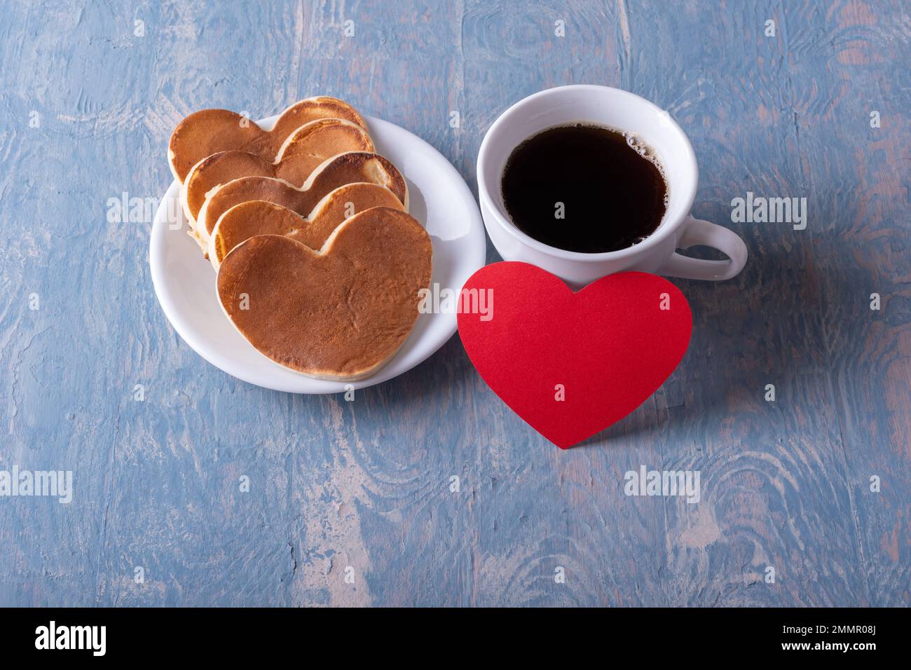Hausgemachte herzförmige Pfannkuchen auf weißem Teller, eine Tasse mit Kaffee oder Kakao und ein rotes, rotes Herz mit blauem, stilvollem Holzhintergrund, Draufsicht. Stockfoto