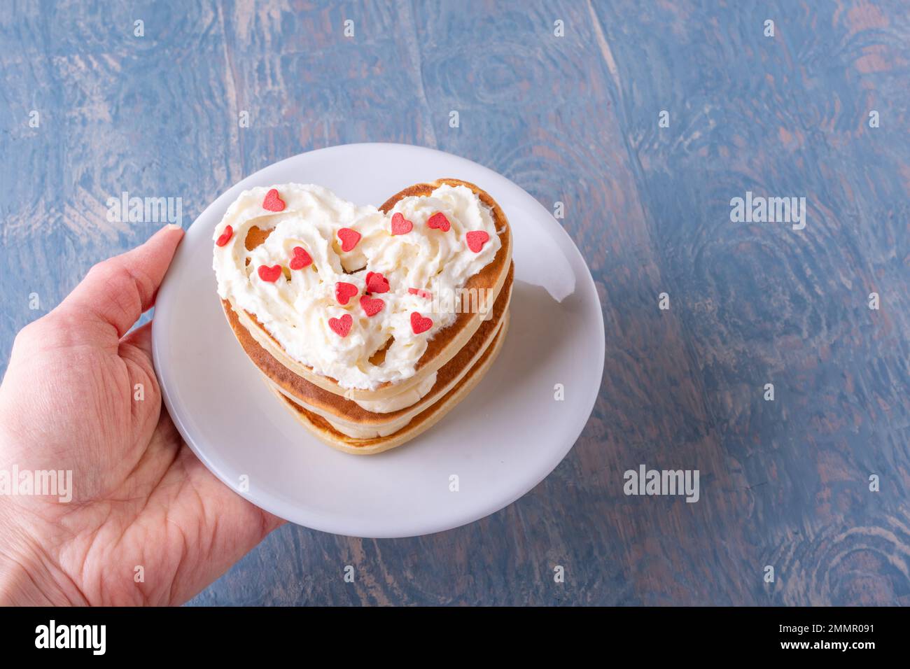 Kreatives Frühstück am Valentinstag. Weibliche Hand hält einen weißen Teller mit einem Stapel hausgemachter herzförmiger Pfannkuchen, dekoriert mit weißer Creme und Stockfoto