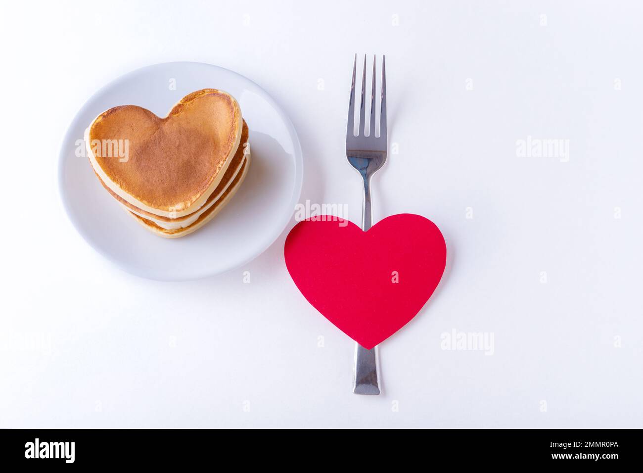Hausgemachte herzförmige Pfannkuchen auf weißem Teller, Gabel und rotes, rotes Herz auf weißem Hintergrund, Draufsicht, Kopierbereich. Postkarte für Valentinstag Stockfoto