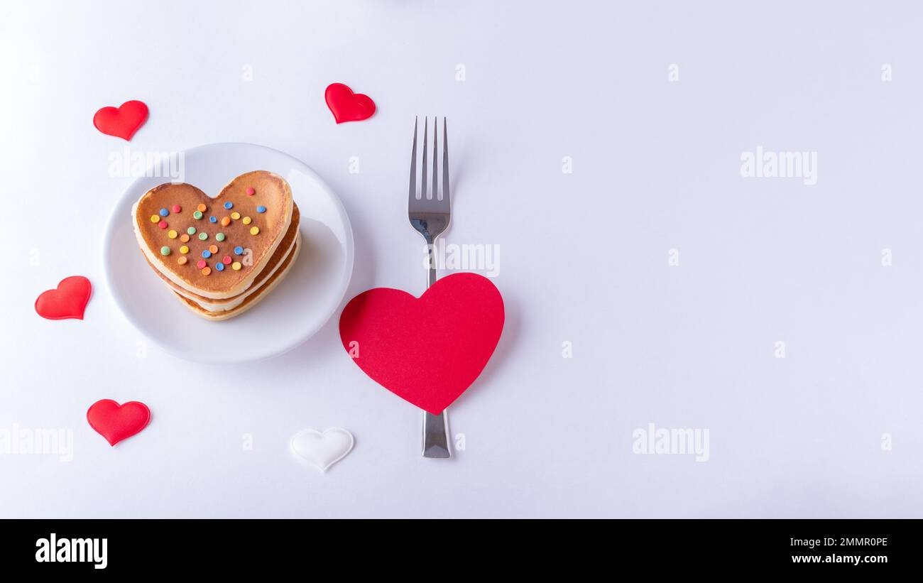 Hausgemachte herzförmige Pfannkuchen, dekoriert mit Zuckerbonbons auf einem weißen Teller, einer Gabel und einer roten Herzform auf weißem Hintergrund mit Herzen, Draufsicht, Stockfoto