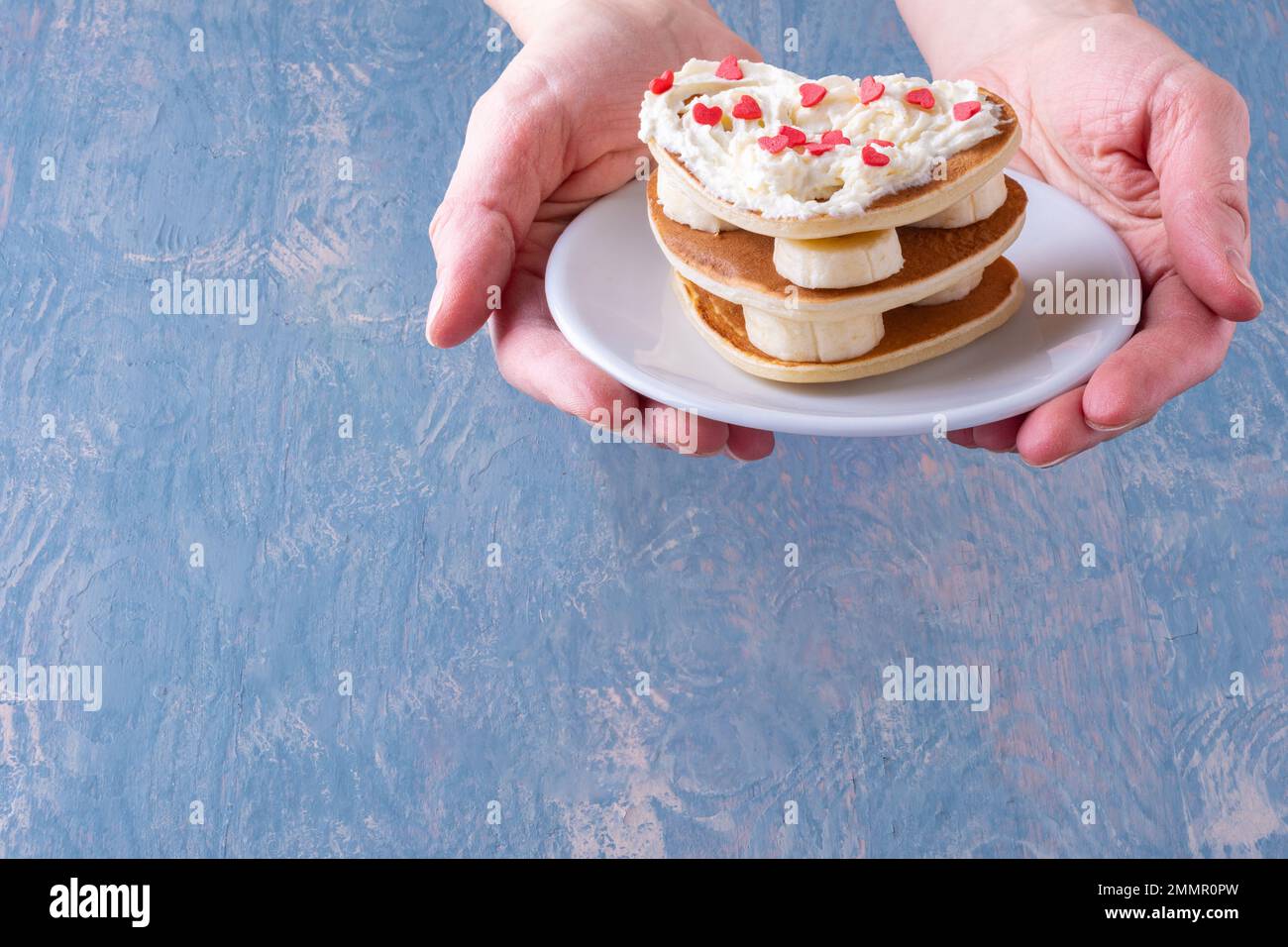 Kreatives Frühstück im Bett. Weibliche Hand hält einen weißen Teller mit einem Stapel hausgemachter herzförmiger Pfannkuchen, dekoriert mit weißer Creme und roten Herzen Stockfoto