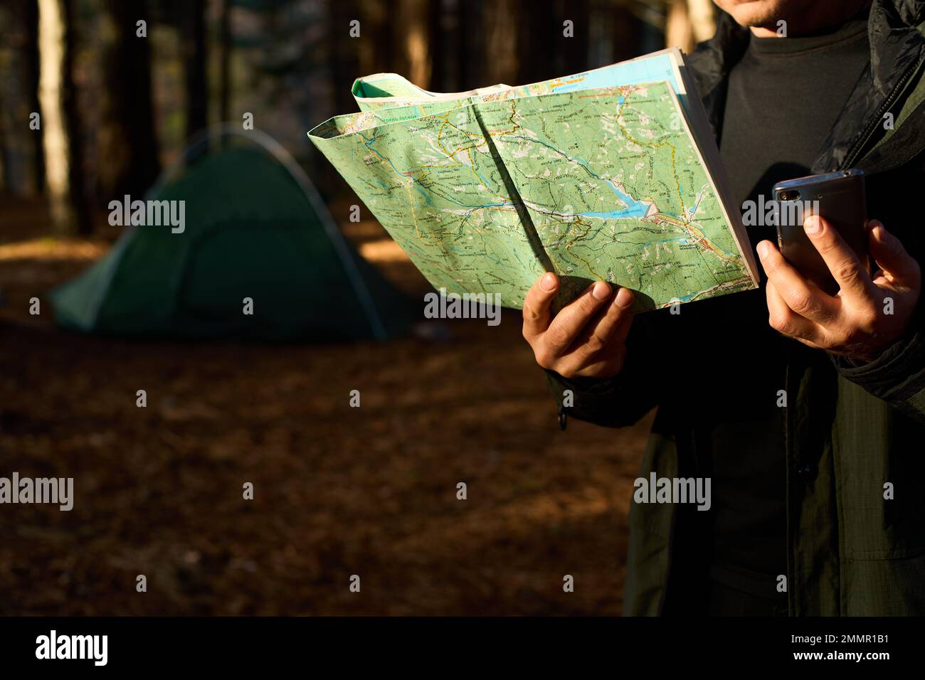Erwachsener Wanderer mit Rucksack, der Handy und Karte benutzt, im Wald in der Nähe seines Zelts. Wandern älterer Mann in der herbstlichen Natur mit Karte im Freien neben einem Stockfoto