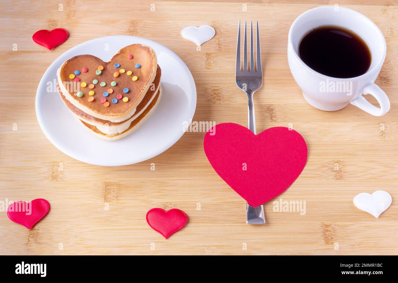 Hausgemachte herzförmige Pfannkuchen mit Zuckerdekoration auf einem weißen Teller, einer Gabel in roter Herzform, einer Tasse mit Kaffee oder Kakao auf Holzhintergrund Stockfoto