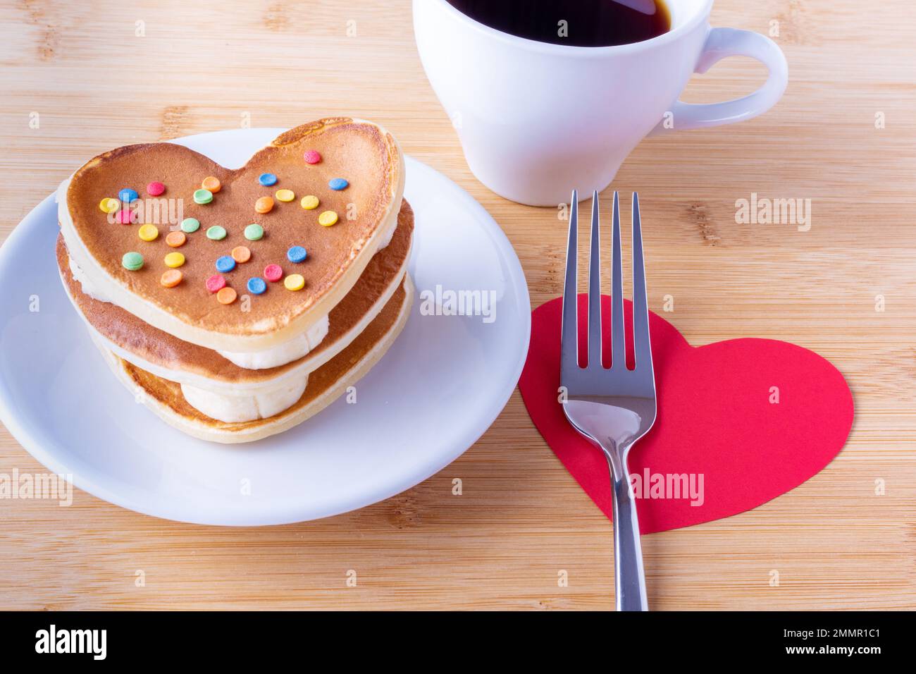 Hausgemachte herzförmige Pfannkuchen mit Zuckerdekoration auf einem weißen Teller, einer Gabel auf rotem Herzen, einer Tasse mit Kaffee oder Kakao, Nahaufnahme. Frühstück für Stockfoto