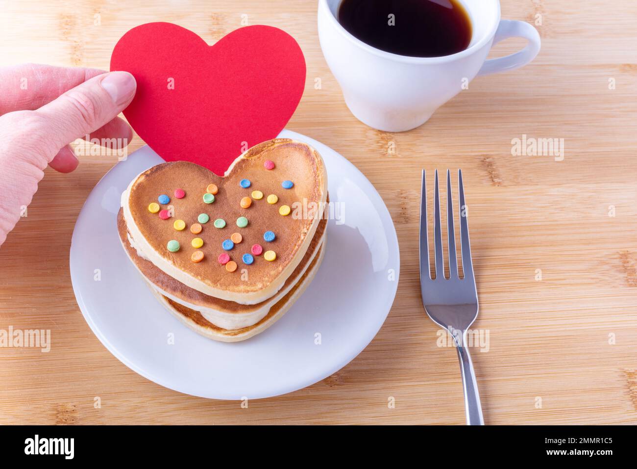 Eine Hand hält eine rote, leere Herzform neben hausgemachten herzförmigen Pfannkuchen mit Zuckerdekoration auf einem weißen Teller, einer Gabel, einem Becher mit Kaffee oder Kakao, clos Stockfoto