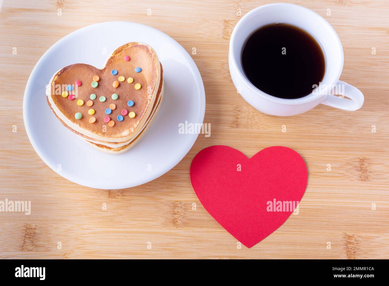 Kreatives Frühstück zum Valentinstag. Hausgemachte herzförmige Pfannkuchen mit Zuckerdekoration auf weißem Teller, Gabel, Becher mit Kaffee oder Kakao und rotem Saum Stockfoto