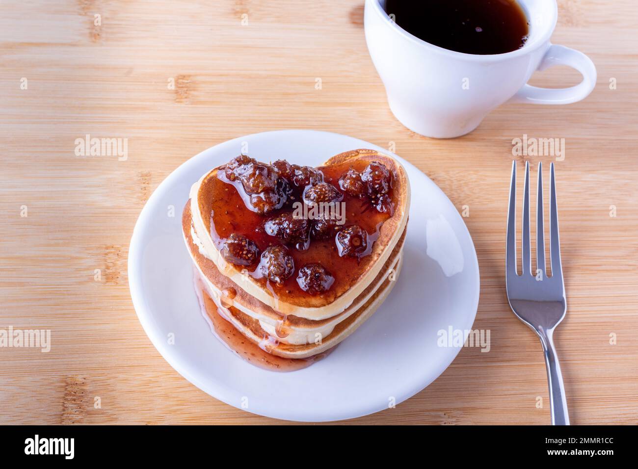 Kreatives Frühstück im Bett. Hausgemachte herzförmige Pfannkuchen mit Beerenmarmelade auf weißem Teller, Gabel und Tasse mit Kaffee oder Kakao auf Holzhintergrund. P Stockfoto