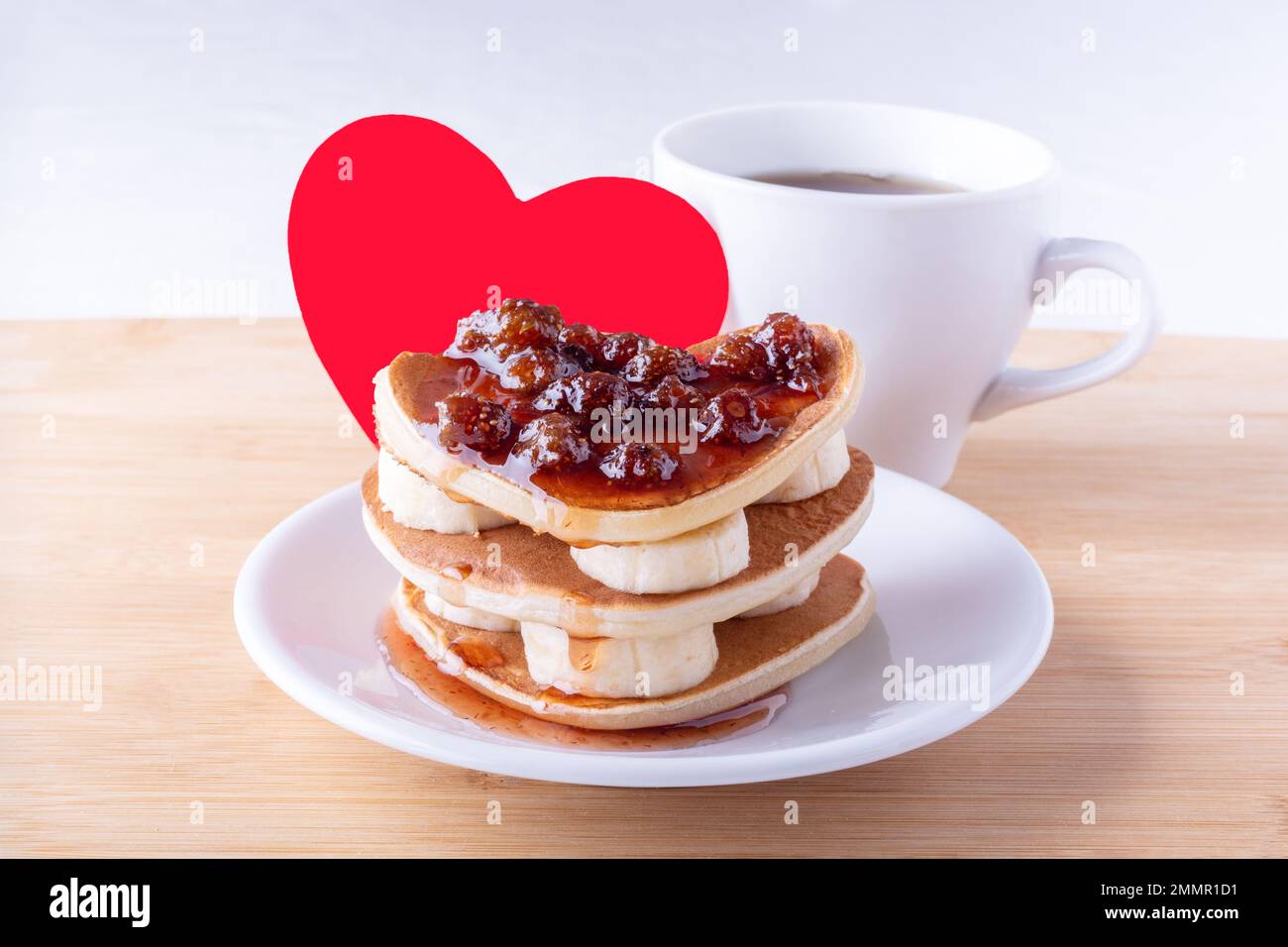 Kreatives Frühstück zum Valentinstag, Muttertag, Geburtstag. Hausgemachte herzförmige Pfannkuchen mit Beerenmarmelade und Bananen auf weißem Teller, Gabel, Becher Stockfoto