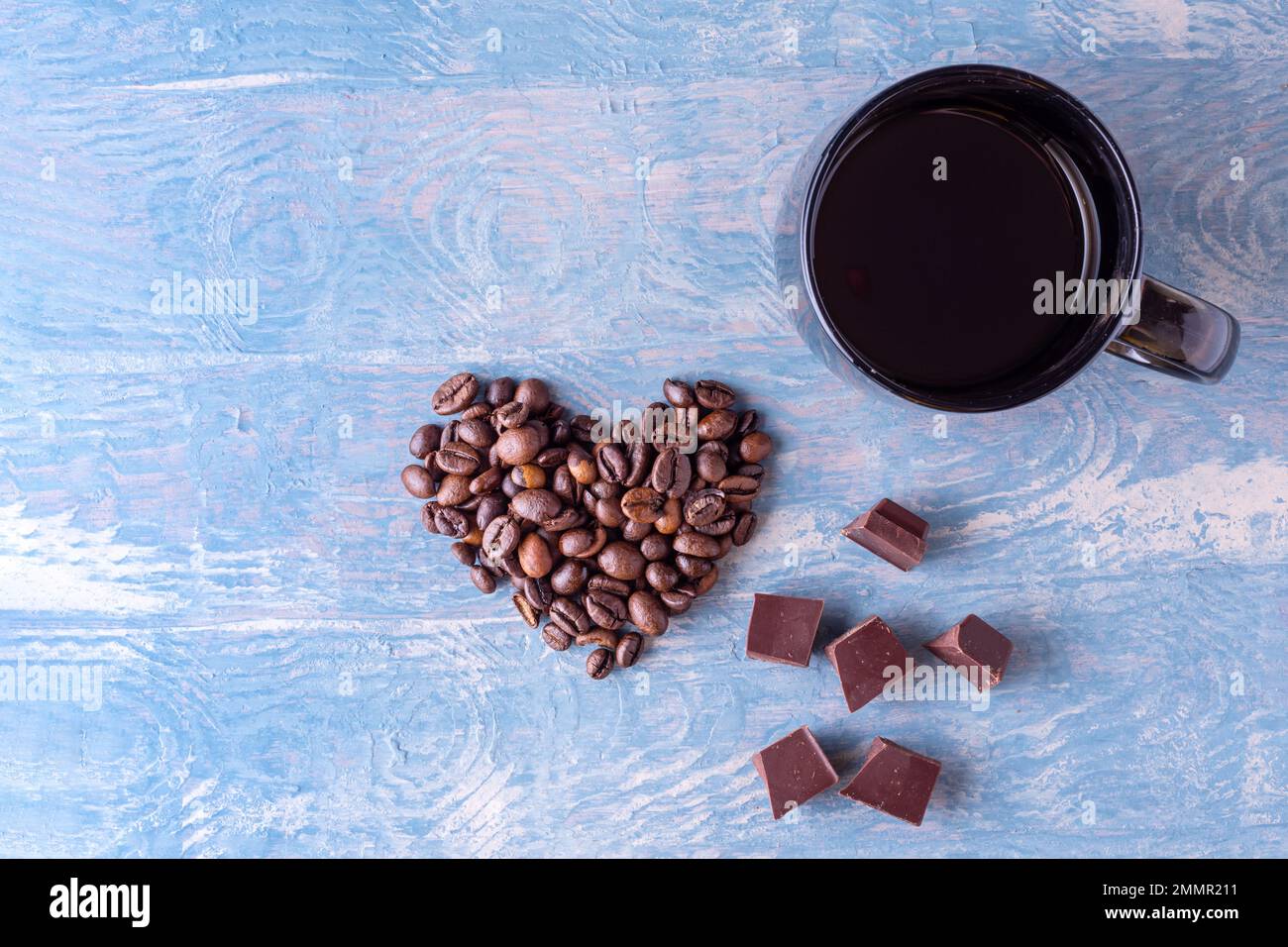 Symbol der Liebe zu Kaffee und Schokolade. Herzform aus Kaffeebohnen, einer großen dunklen Tasse Kaffee und Stücken schwarzer dunkler Schokolade auf einem blauen W. Stockfoto
