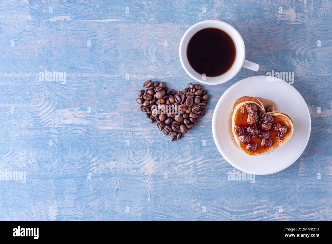Frühstück für deine Geliebte. Hausgemachte herzförmige Pfannkuchen mit Beerenmarmelade, herzförmig aus Kaffeebohnen und einer weißen Tasse heißem Kaffee auf einem blauen Stockfoto