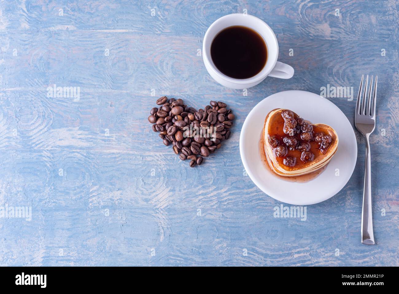Frühstück für Dad. Hausgemachte herzförmige Pfannkuchen mit Beerenmarmelade, herzförmig aus Kaffeebohnen, weißer Tasse Kaffee und Gabel auf blauem Holztisch Stockfoto