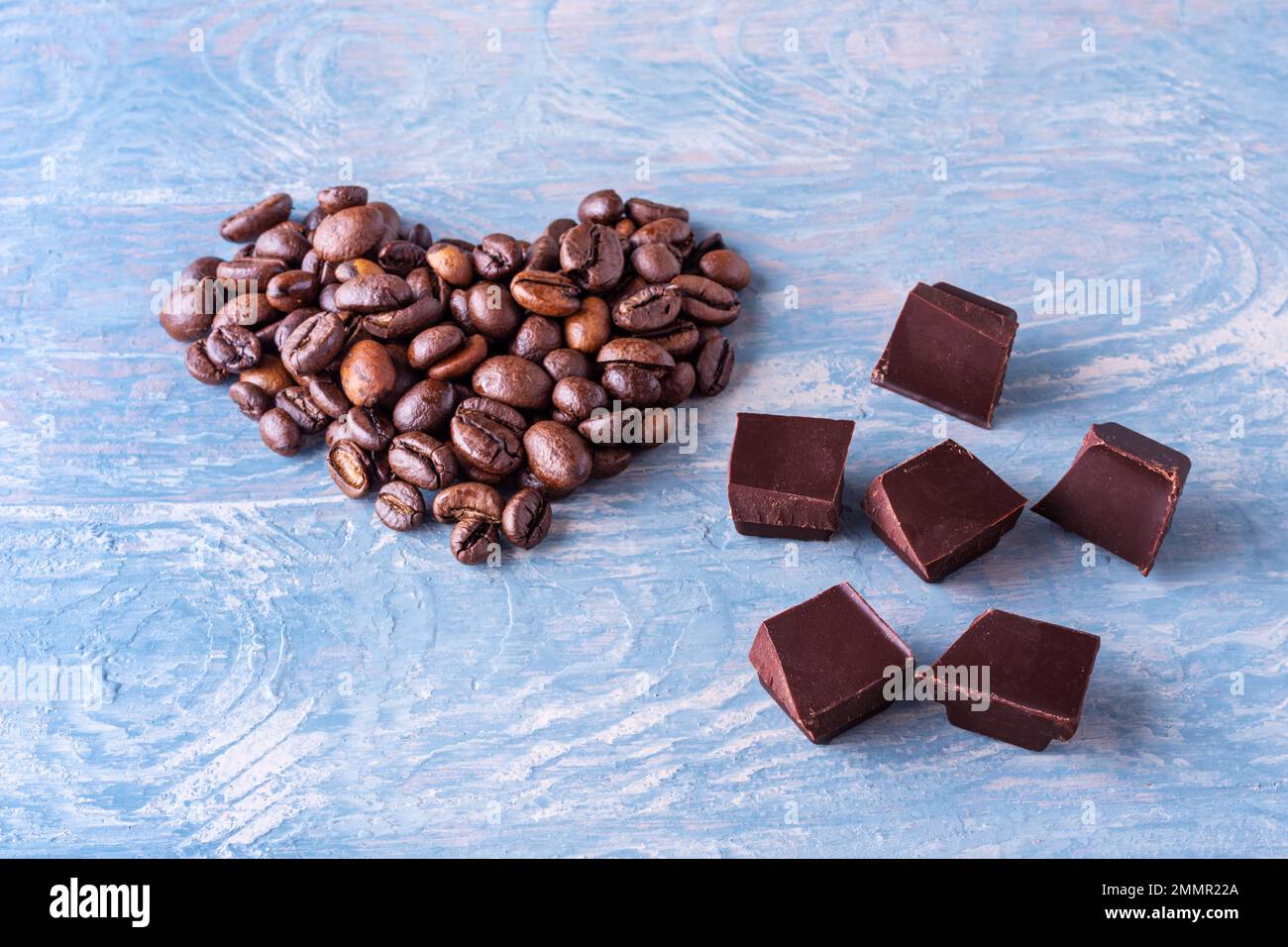 Symbol der Liebe zu Kaffee und Schokolade. Herzform aus Kaffeebohnen und Stücken schwarzer bitterer Schokolade auf blauem Holzhintergrund, Nahaufnahme Stockfoto