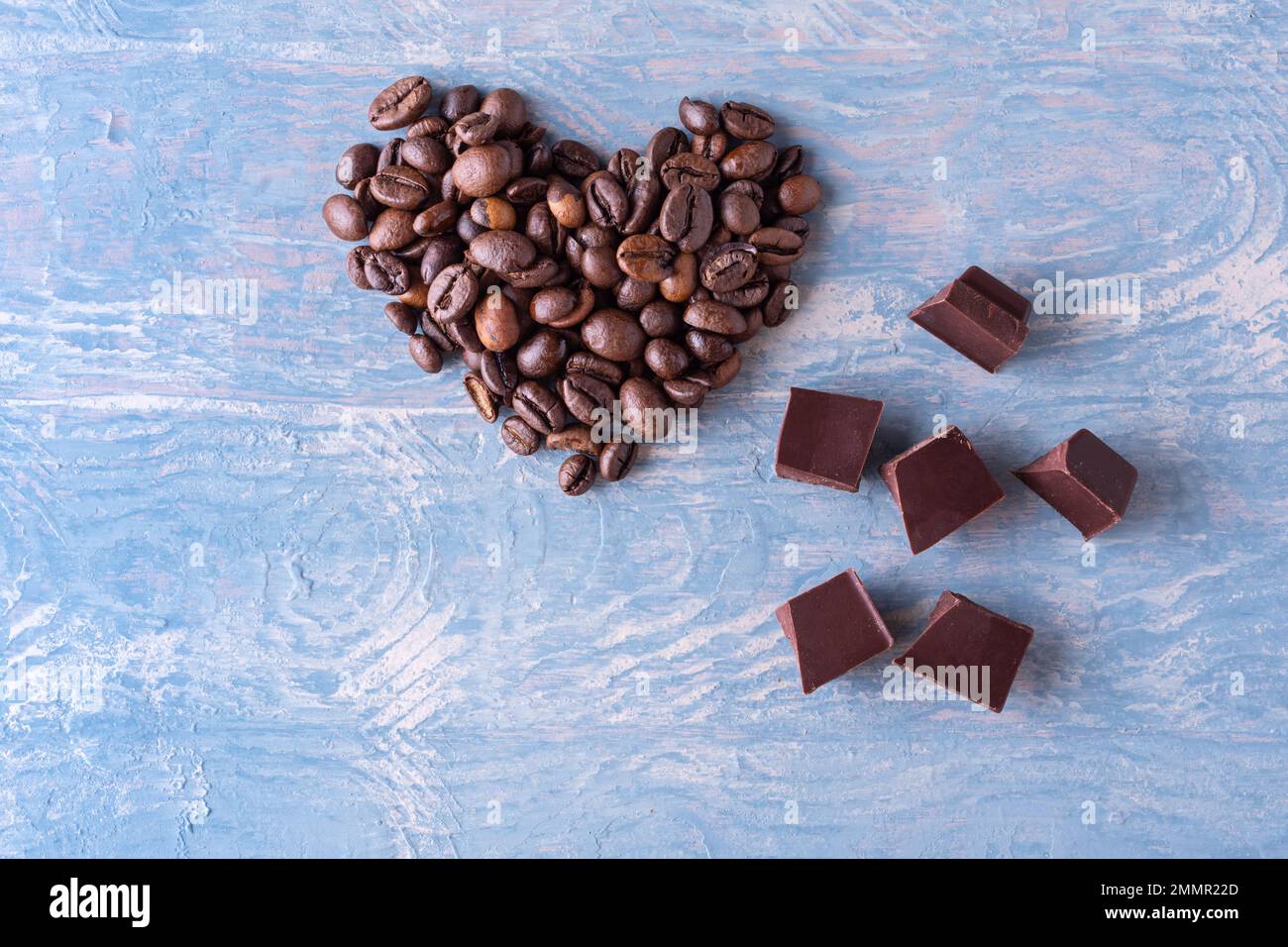 Symbol der Liebe zu Kaffee und Schokolade. Herzform aus Kaffeebohnen und Stücken schwarzer bitterer Schokolade auf blauem Holzhintergrund, Nahaufnahme Stockfoto