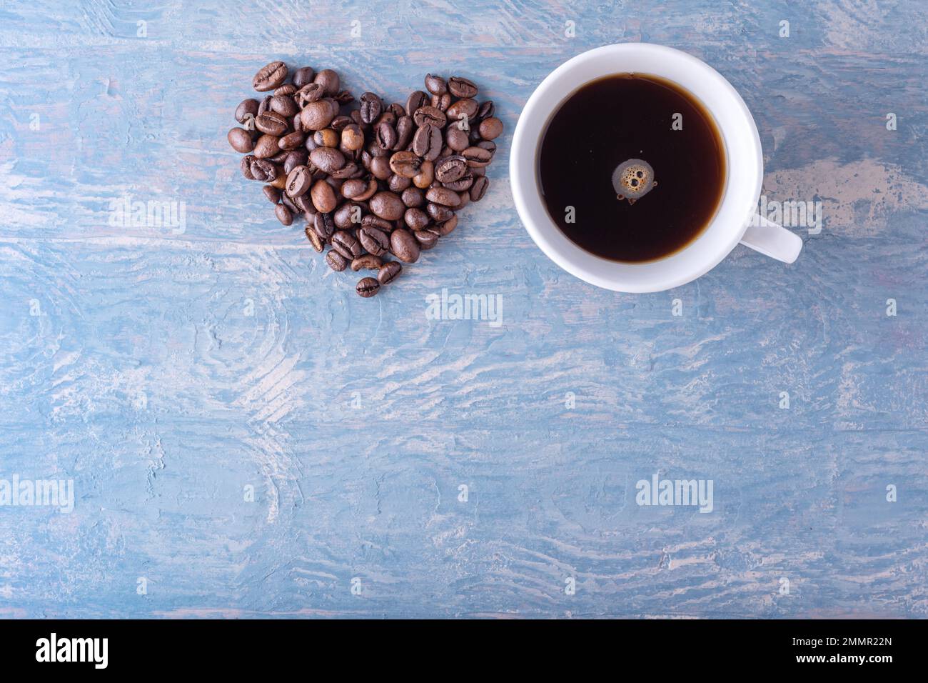 Herzform aus Kaffeebohnen und weißer Kaffeetasse auf blauem, stilvollem Holzhintergrund, Nahaufnahme, Draufsicht, flach liegend. Symbol der Liebe zum Kaffee. Stockfoto