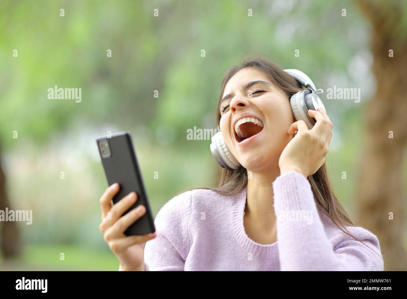 Ein glücklicher Teenager, der in einem Park singt und Musik mit Kopfhörern und Telefon hört Stockfoto