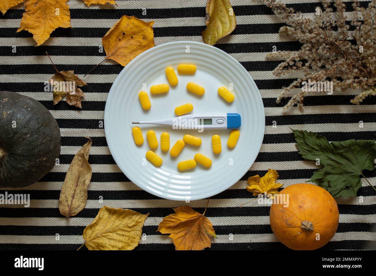 Ein elektronisches Thermometer liegt neben gelben Pillen auf einem Teller auf dem Tisch in der Küche mit Blättern und Kürbis in der Nähe, Medizin und Apotheke heilen Stockfoto