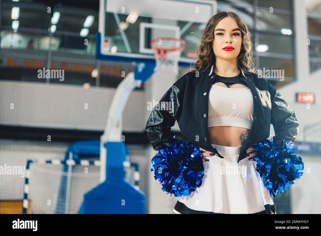 Horizontaler mittlerer Schuss einer Cheerleaderin schwarz-weißer Uniform, die sich mit blau glänzenden Bommeln posiert. Basketballfeld verschwommen im Hintergrund. Hochwertiges Foto Stockfoto