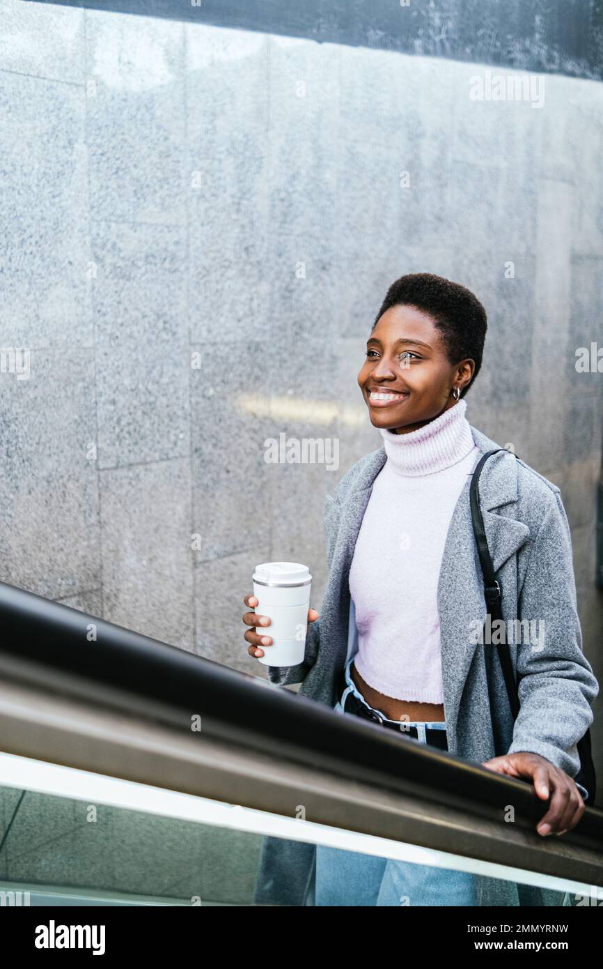 Hochwinkel einer optimistischen afroamerikanischen Frau in stilvoller Oberbekleidung. Sie hält eine Tasse Kaffee ohne Abfall und sie lächelt und fährt tagsüber in der Stadt Treppen aus der Untergrundbahn Stockfoto