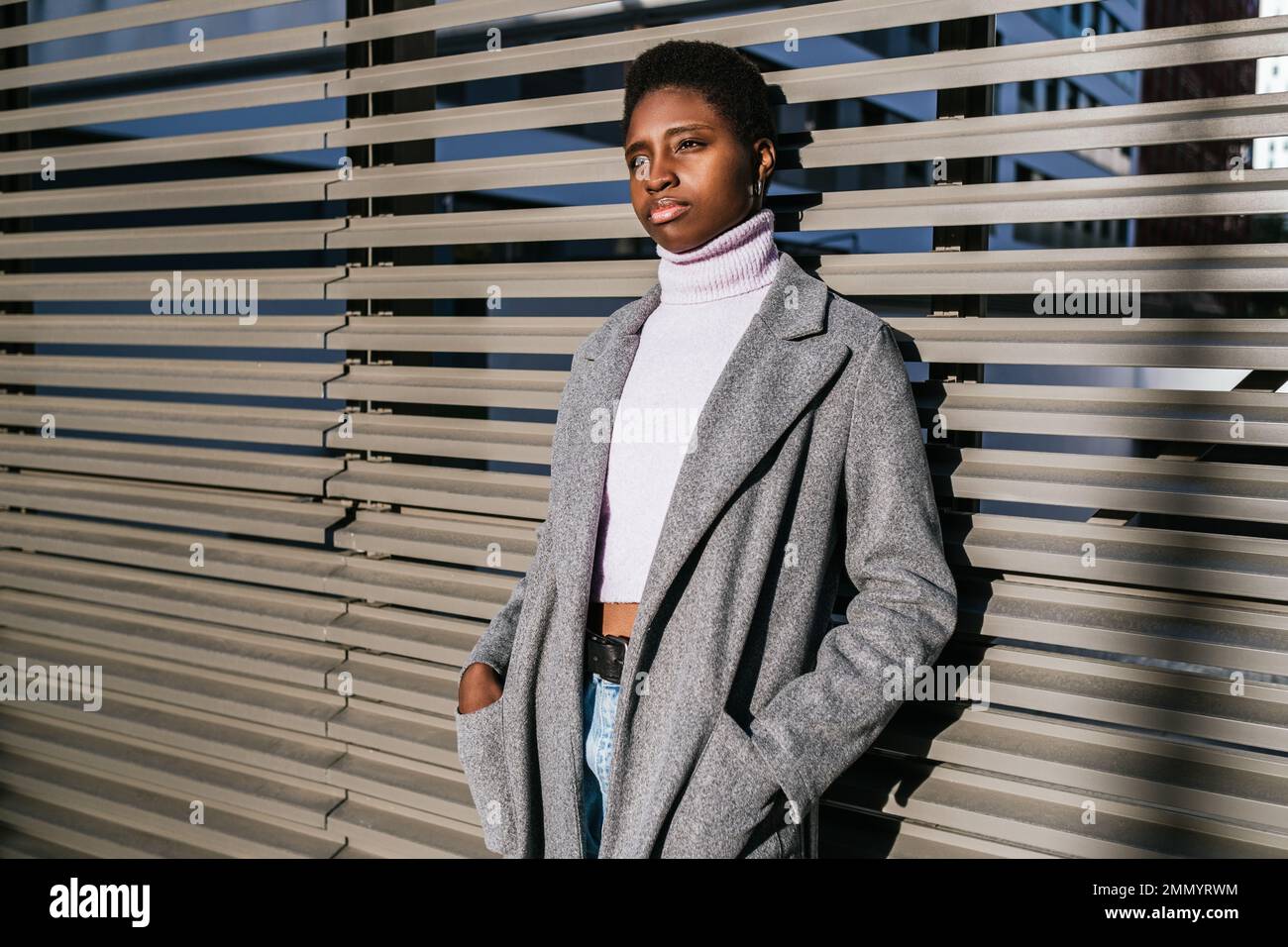 Eine unglückliche afroamerikanische Frau in einem stilvollen Mantel und Pullover, die Hände in den Taschen hält und wegschaut, während sie sich an die sonnenbeleuchtete Wand der Stadtstraße lehnt Stockfoto