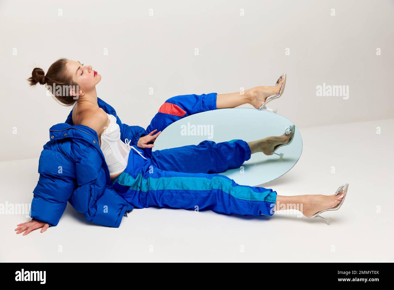 Porträt einer schönen Frau in sportlicher Retro-Hose und Daunenjacke mit Spiegel vor hellem Hintergrund. Das Konzept von seltsamen Menschen, Mode Stockfoto