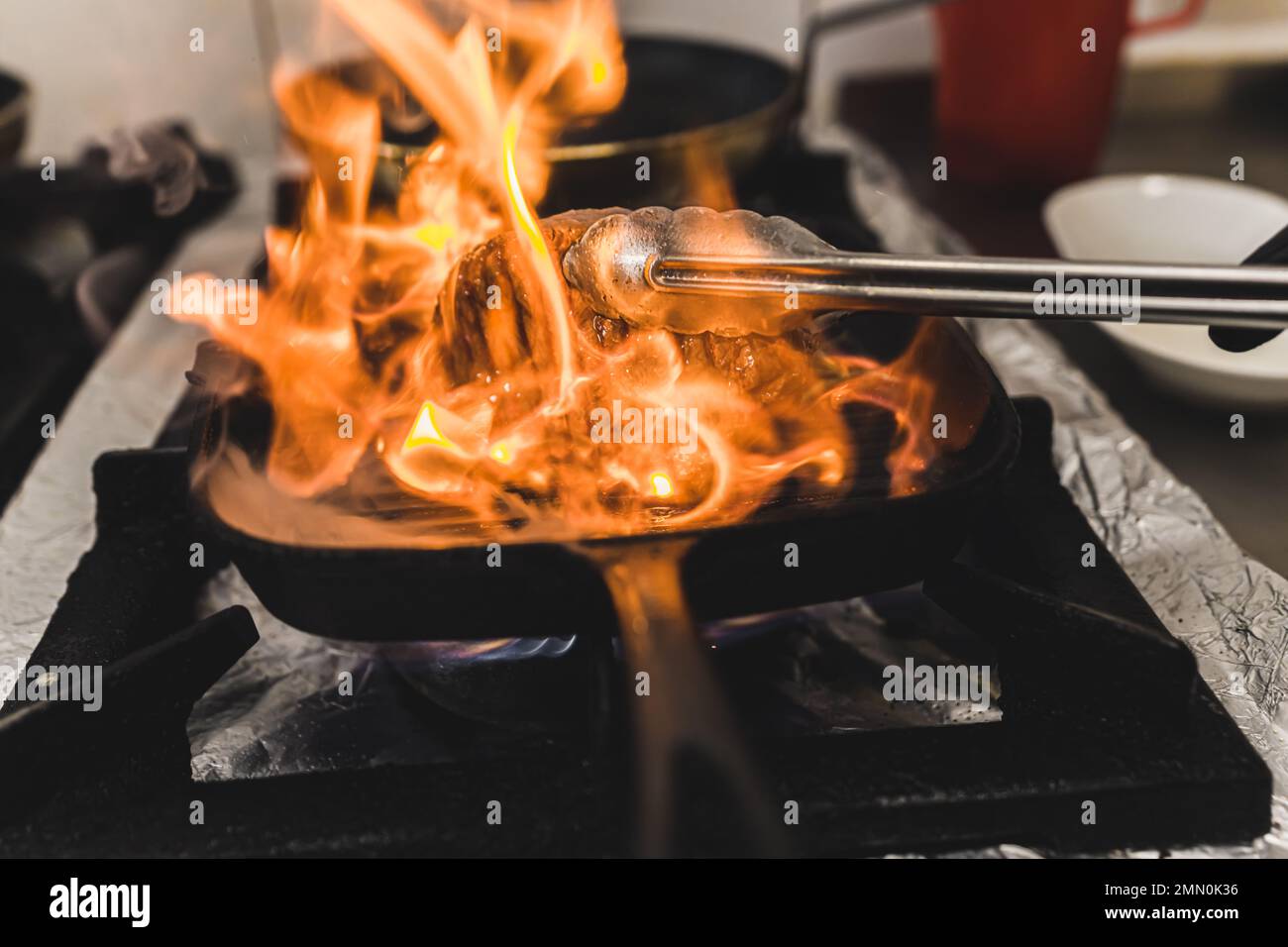 Eine unbekannte Person, die Streifenfilet-Steak mit Zange umwirft. Flammende Grillpfanne. Zubereitung von Speisen im Restaurant. Horizontale Indoor-Aufnahme. Hochwertiges Foto Stockfoto