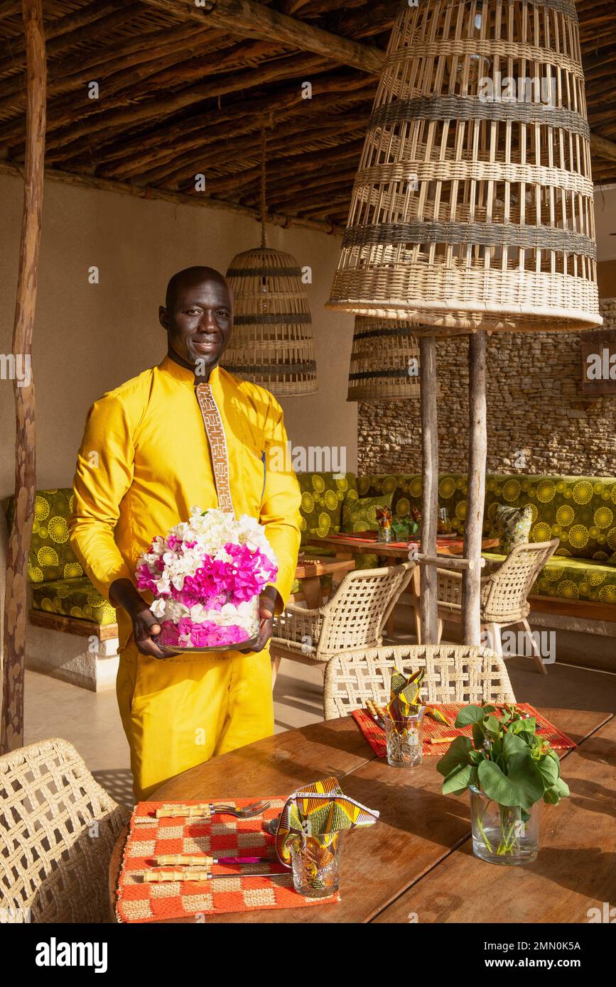 Senegal, Saloum Delta, UNESCO-Weltkulturerbe, Palmarin, Yokan Lodge, Kellner in elegantem gelben Outfit mit einem Tablett mit Blumen in dem rustikalen schicken Restaurant aus natürlichen Materialien Stockfoto