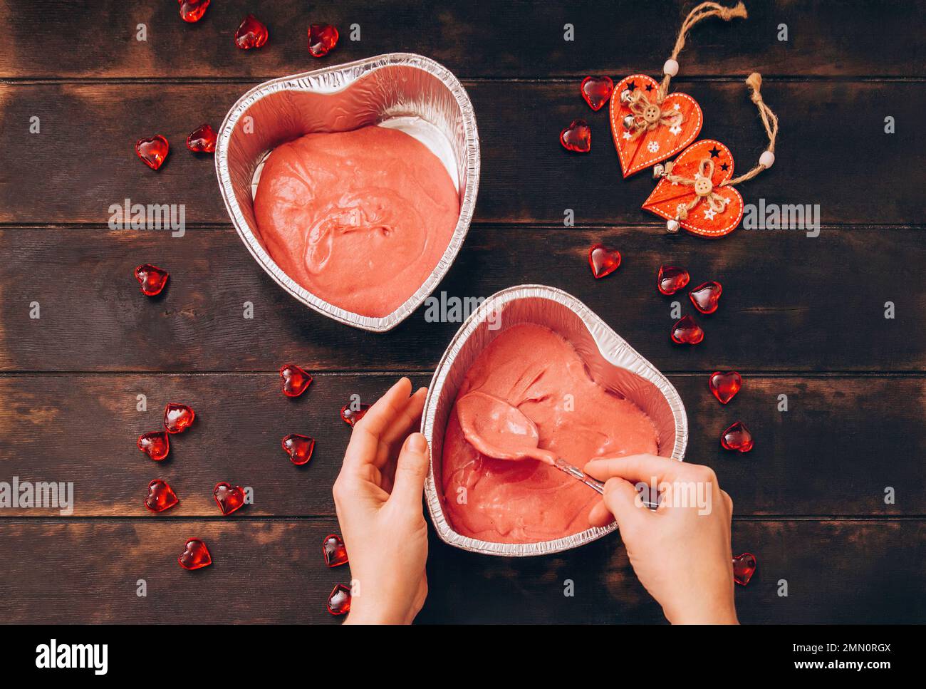 Frauenhände mit einem Löffel geben den Teig in Form von Folie, um einen Kuchen in Form eines Herzens zu backen. Vorbereitung auf den Valentinstag. Stockfoto