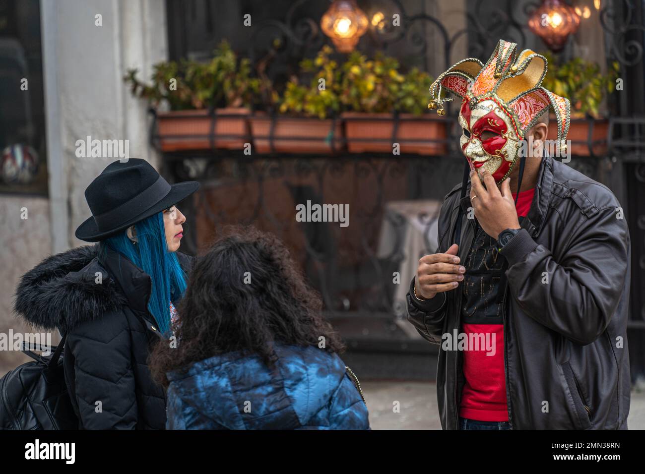Ein Mann mit einer Maske spricht mit zwei Frauen, von denen eine mit dem blauen Haar auf der Straße während des Venedig-Jahrmarkts ist Stockfoto