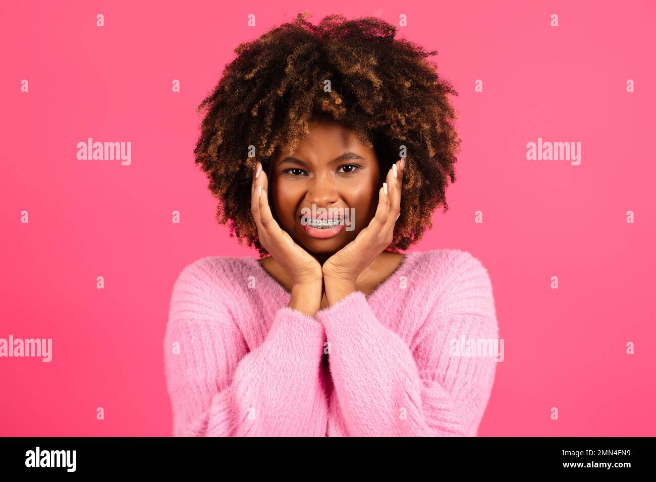 Traurige, unzufriedene, lächelnde junge, schwarze, geschweifte Dame mit einer Zahnspange in lässiger Form presst Hände auf die Wangen und leidet an Zahnschmerzen Stockfoto